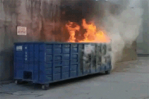 Large blue dumpster on fire outside a building A dumpster is engulfed in flames. Fire and smoke are visible.
