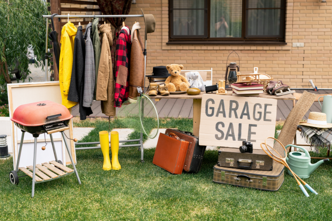 A garage sale display on a lawn showcases clothes on a rack, a teddy bear, red grill, yellow boots, suitcases, and vintage items—an inspiring scene for anyone interested in garage sales or seeking the ultimate guide to decluttering.