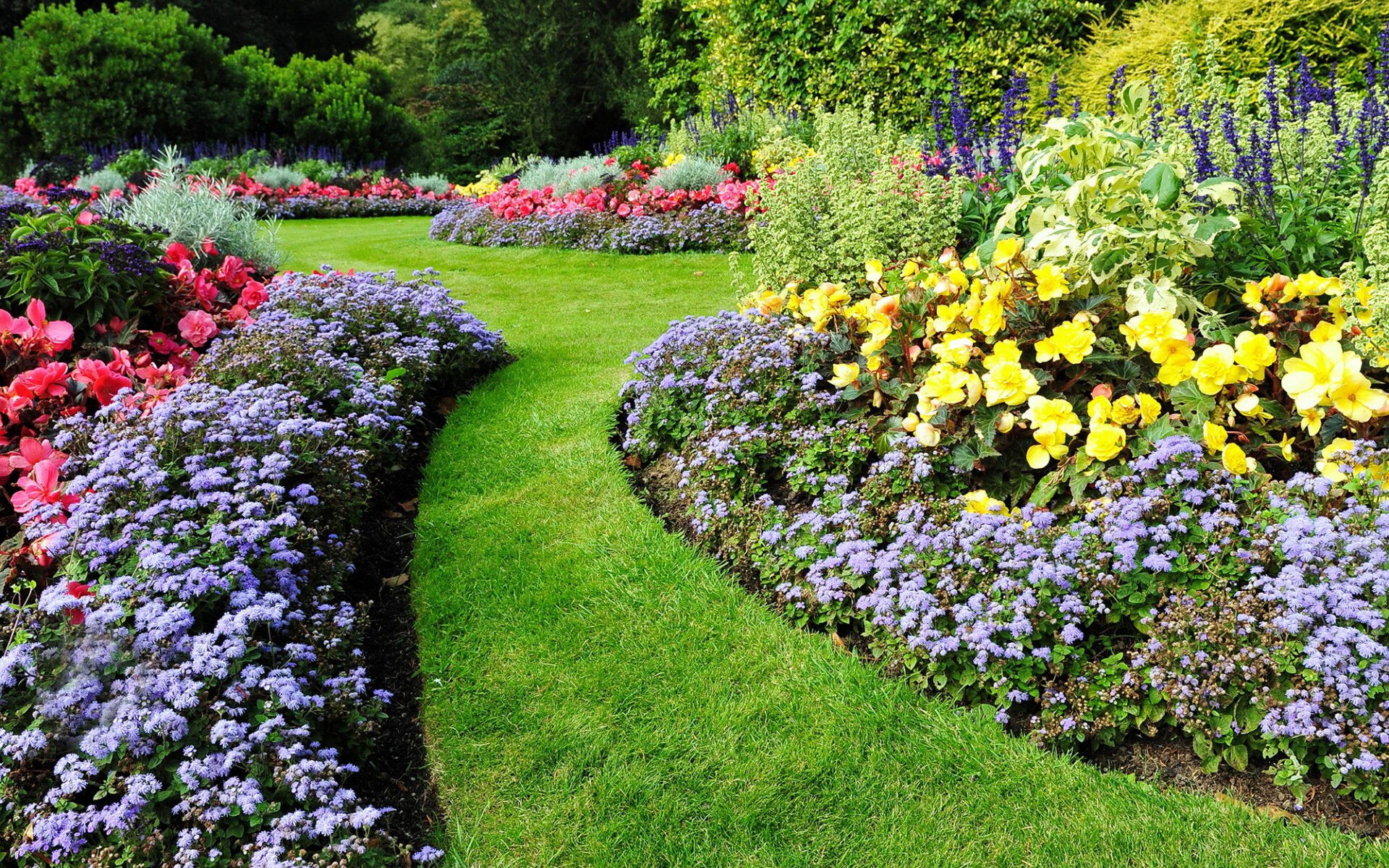 A well-manicured garden path lined with purple, yellow, pink and red flowers. Lush greenery surrounds the garden.