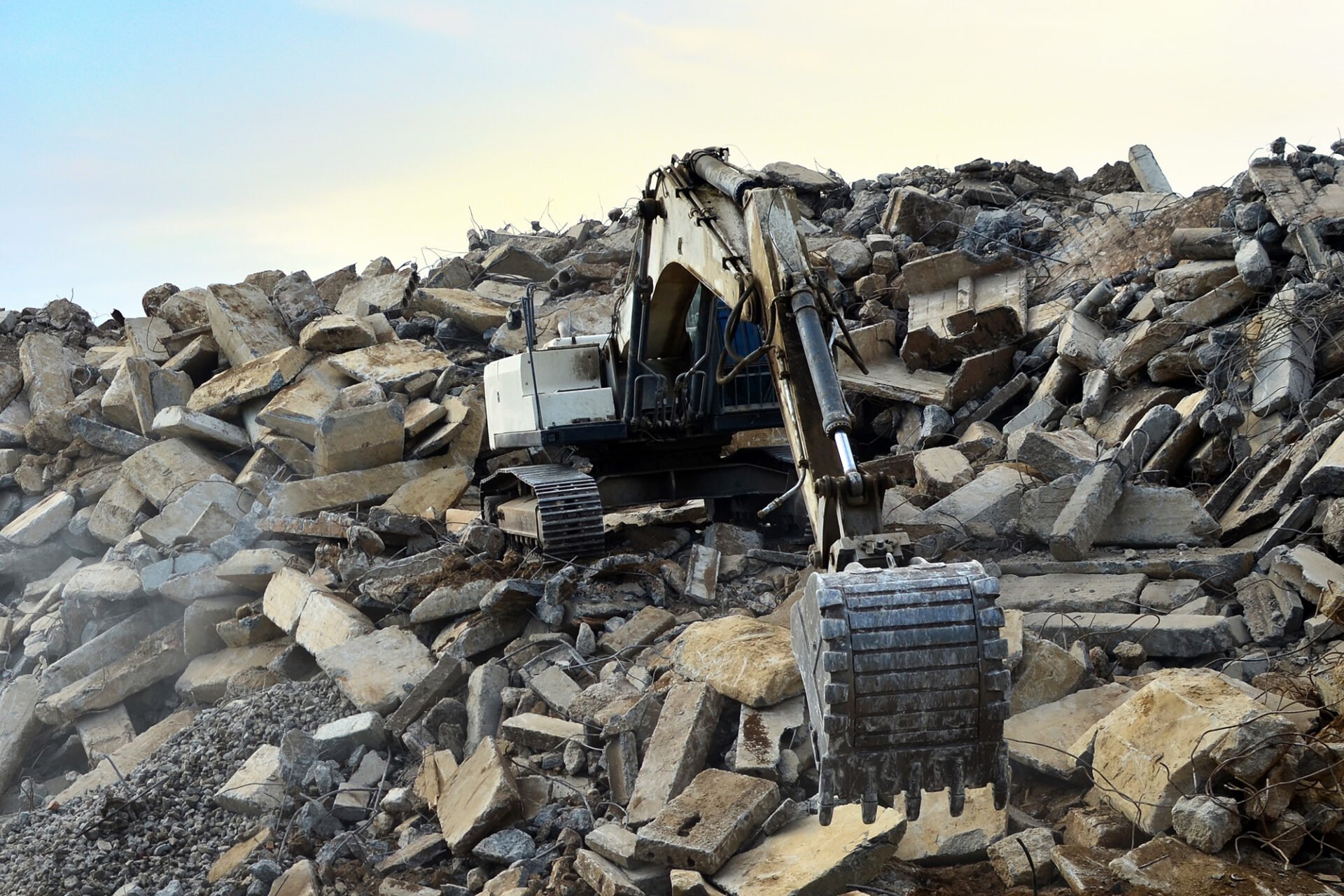 An excavator on a large pile of rubble and debris. The machine is surrounded by broken concrete and rocks.