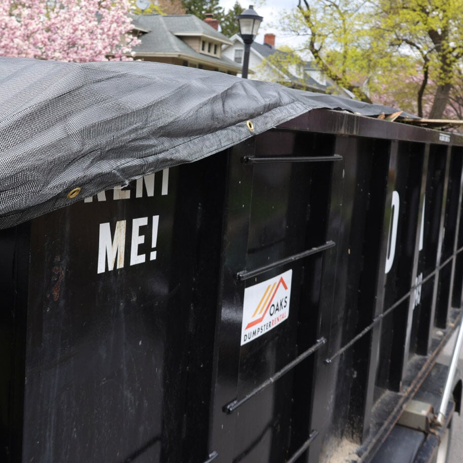 Black dumpster with gray tarp on residential street A black dumpster with a gray tarp on top sits on a residential street.