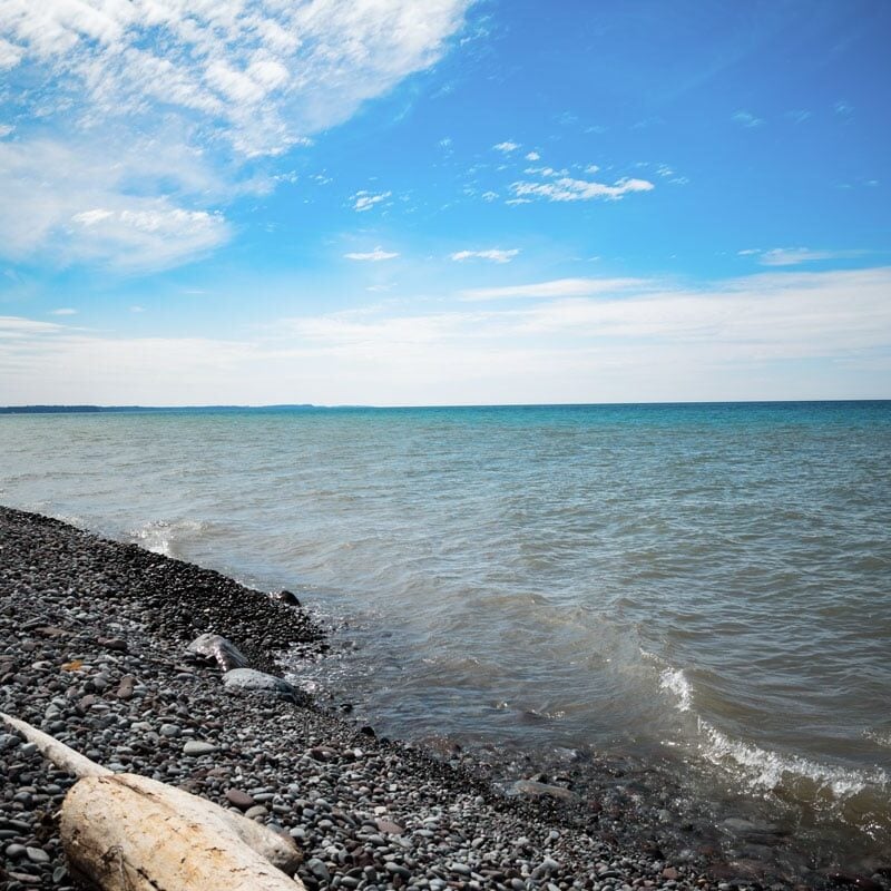 Serene Lake Shore with Pebbles and Driftwood A peaceful lake shore with pebbles and driftwood. Calm water and blue sky with clouds.